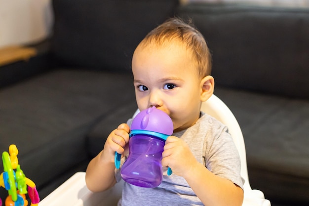 Sweet baby boy sitting in high baby chair and drinking water from baby bottle Baby at home