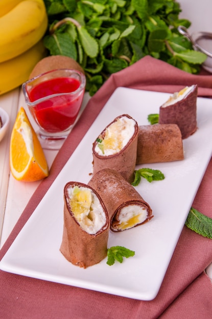 Sweet chocolate rolls from pancakes with cream cheese, fresh fruit and berry sauce on a white wooden table.