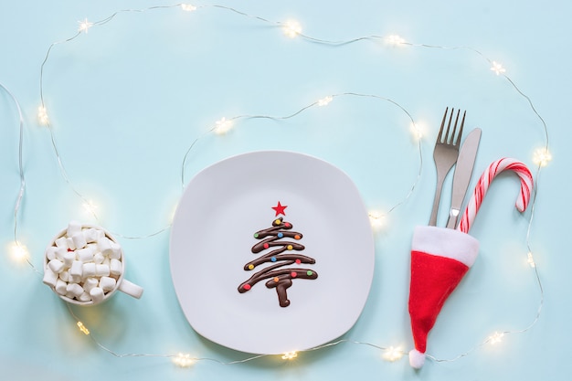 Sweet Christmas tree made of chocolate, cutlery and cup of cocoa marshmallow and light bulbs garland 