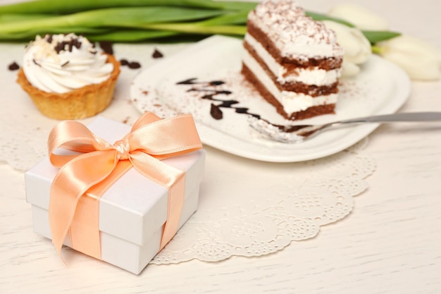 Sweet creamy cake with flowers and present box on wooden table closeup