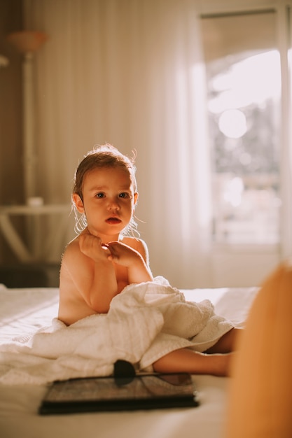 Sweet curly little girl with a towel over her wet body siting in a bedroom after shower