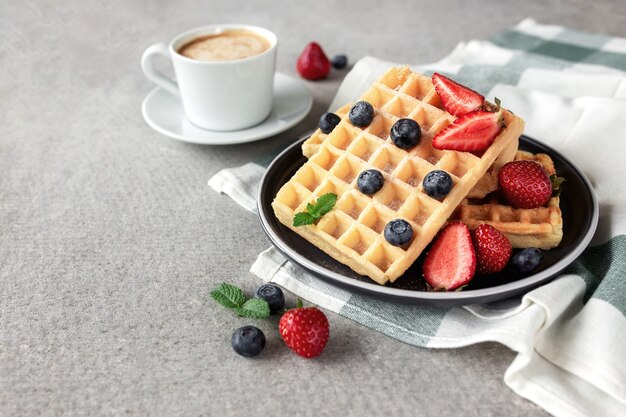 Sweet delicious breakfast with plate of waffles, strawberry and blueberry, mint leaf and cup of coffee on concrete gray background. High quality photo