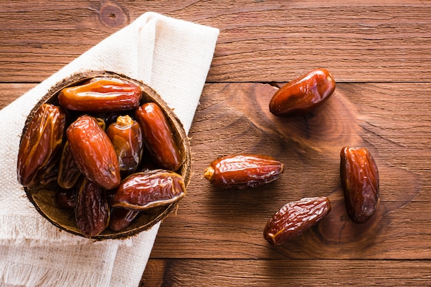 Sweet dried dates fruit in a small bowl on napkin on wood table. Top view