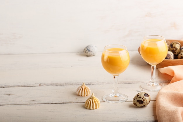 Sweet egg liqueur in glass with quail eggs and meringues on white wooden background. Side view, copy space.