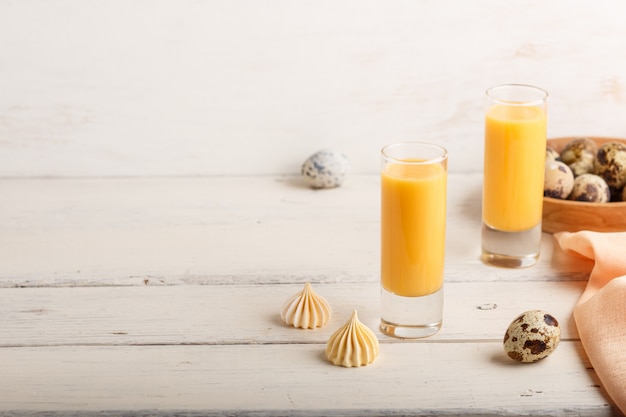 Sweet egg liqueur in glass with quail eggs and meringues on white wooden background. Side view, copy space.