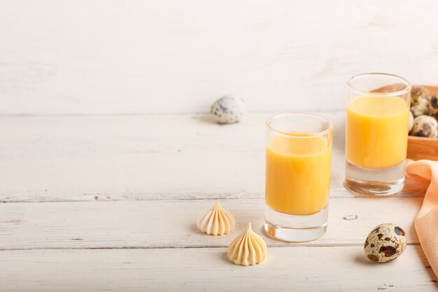 Sweet egg liqueur in glass with quail eggs and meringues on white wooden background. Side view, copy space.