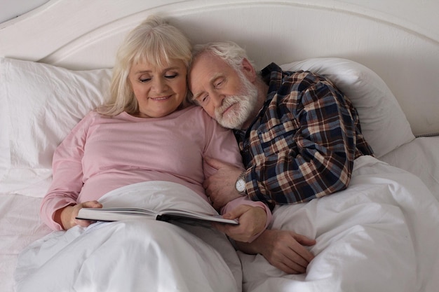 Sweet elderly married couple reading book in bed. Old man resting his head on his wifes shoulder in their bedroom reading magazine.