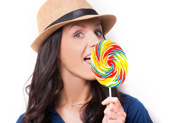 Sweet and extremely big. Surprised young beautiful woman in funky hat licking a big lollipop while standing against white background