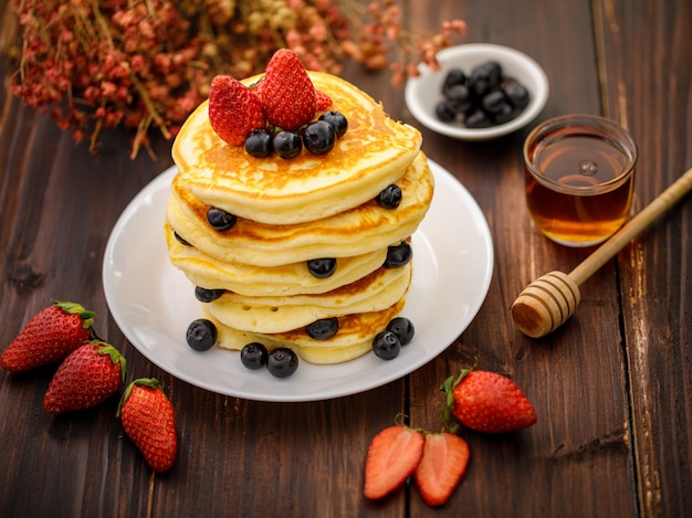 달콤한 음식. 흐릿한 나무 배경에 하얀 접시에 블루베리, 딸기, 꿀을 넣은 맛있는 팬케이크를 쌓으세요.