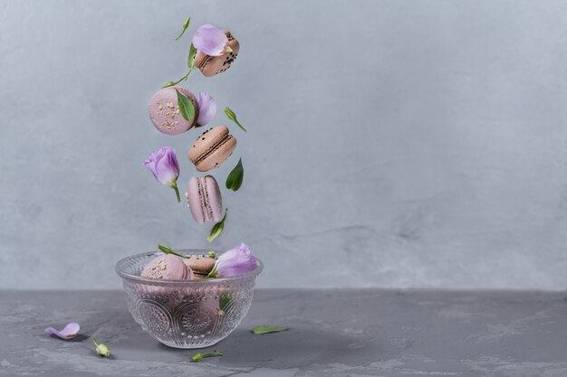 Dolci macarons francesi cadono mescolati con fiori in una ciotola. pastello colorato amaretti volanti biscotti. superficie grigia. concetto di cibo, cucina, al forno e cucina