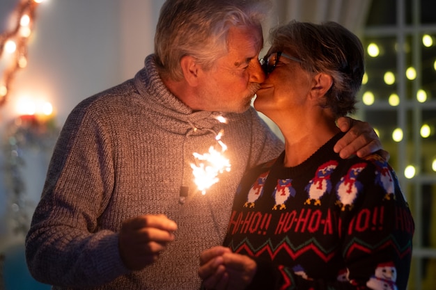 Un dolce bacio tra una coppia anziana di moglie e marito che celebrano il natale con le scintille. luci e albero di natale in sottofondo