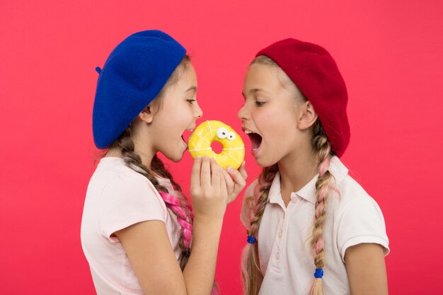 Sweet life. Sweets shop and bakery concept. Kids fans of baked donuts. Share sweet donut. Girls in beret hats hold donut red background. Kids playful girls ready eat donut. Friendship and generosity.