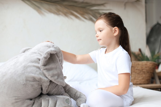 Sweet little girl sitting on her bed at home with toy elephant.