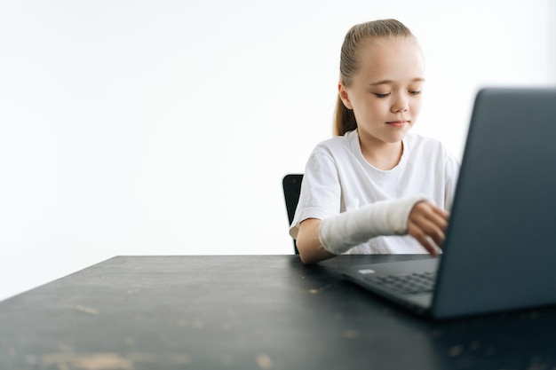 Sweet little girl with broken hand wrapped in white plaster\
bandage typing on laptop computer looking to screen sitting at desk\
in light room