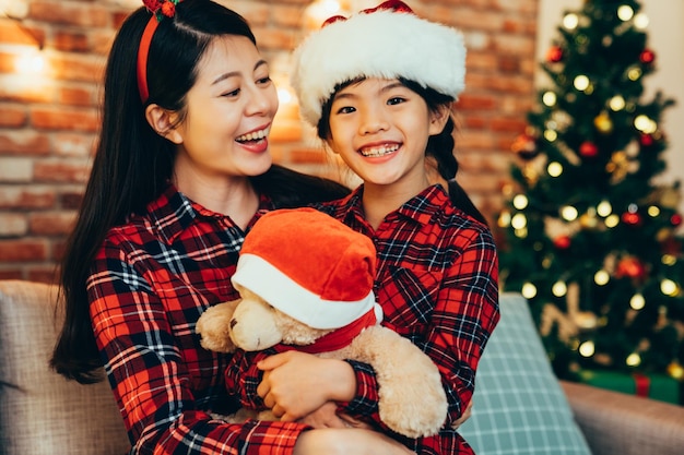sweet lovely family hugging celebrating christmas at home sitting on sofa. smiling little girl with stuffed toy teddy bear wearing white red santa hat. young mom looking cute kid xmas tree background