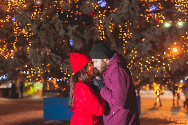 Sweet recently married couple posing near fir tree with Christmas decoration, standing closely face to face and holding hands. Winter holidays concept