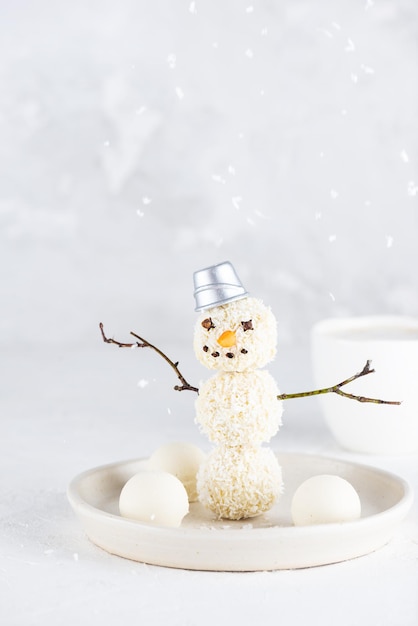 A sweet snowman made of truffles with falling coconut flakes on a light background