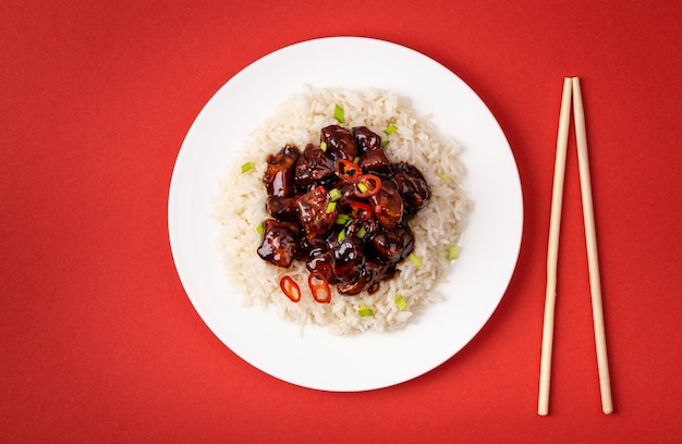 甘酸っぱい肉を白い皿にご飯と粘り気のあるソースで炒め、木の箸、伝統的な中華料理。赤の背景にアジアンディナーまたはランチ、クローズアップ、上面図