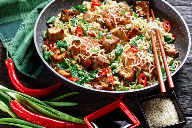 Sweet and sour tofu, chicken thighs, chinese wok noodles, wilted kale, sesame seeds, red chili, green onion, served on a frying pan with chopsticks on a dark wooden background, top view, close-up