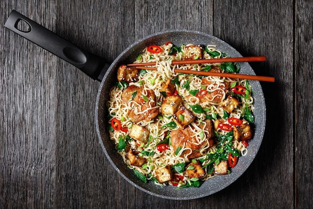 Sweet and sour tofu, chicken thighs, chinese wok noodles, wilted kale, sesame seeds, red chili, green onion, served on a frying pan with chopsticks on a dark wooden background, top view, close-up