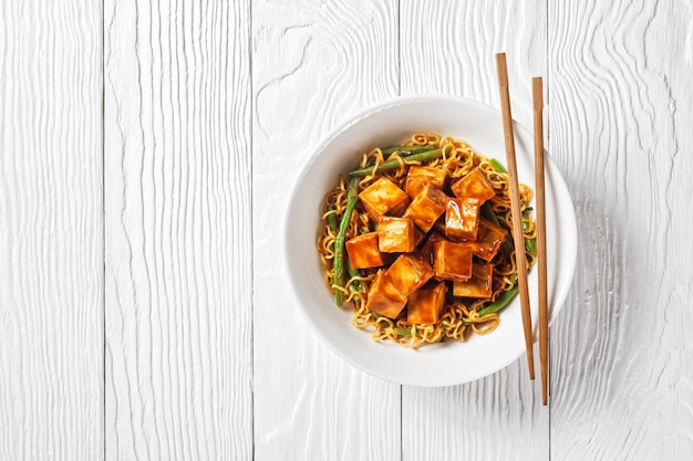 Sweet and spicy asian noodle with hoisin baked crunchy tofu and  green bean  in a white bowl with chopsticks horizontal view from above flat lay free space