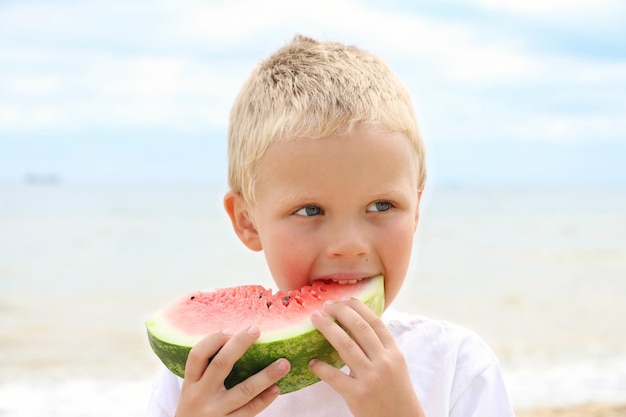 Sweet sun tanned blonde kid on the coast holding juicy slice of the watermelon.