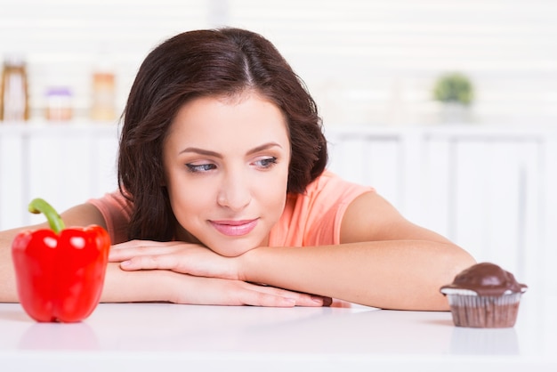 Sweet temptation. Thoughtful young woman choosing what to eat while leaning at the kitchen table with chocolate muffin and fresh pepper laying on it