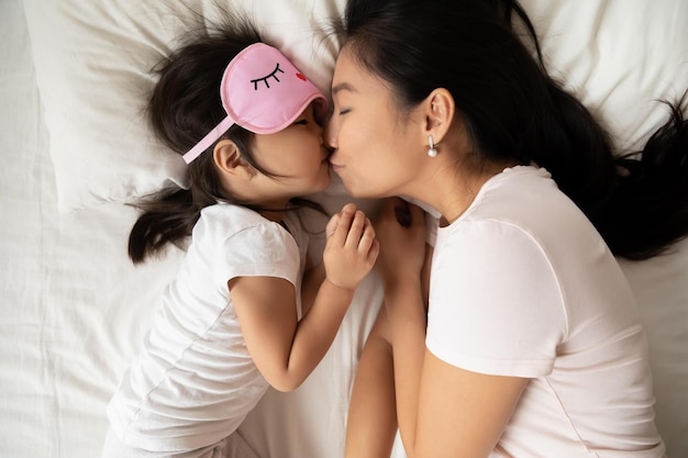 Sweet tender moment of mom kiss child close up top view Loving mum puts child to healthy day night sleep asian family lying in bed kid girl wears pink sleeping eyemask Love health care concept