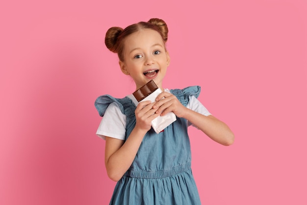 Милая маленькая девочка-сладкоежка кусает плитку шоколада и улыбается в камеру