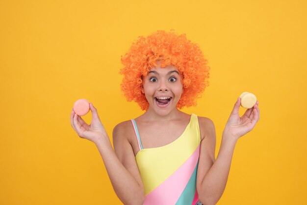 Sweet tooth. funny kid in clown wig hold french macaron. amazed child wear swimsuit. teen dental care. girl with orange hair and french cakes. summer fun. macaroon cookie. happy childhood.