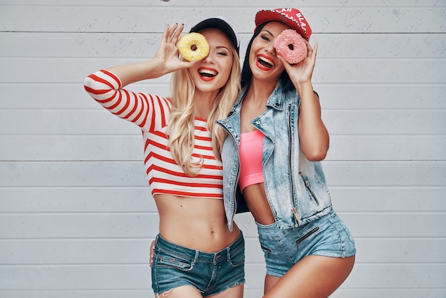 Sweetie beauties. Two playful young women holding donuts against their eyes and smiling while standing against the garage door