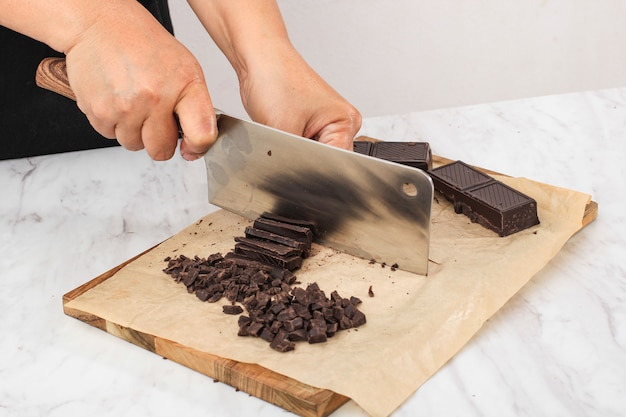 Foto dolci pasticceria e culinaria panificio concetto, mani femminili con coltello da cucina tritare la barra di cioccolato a trucioli su tavola di legno sfondo bianco