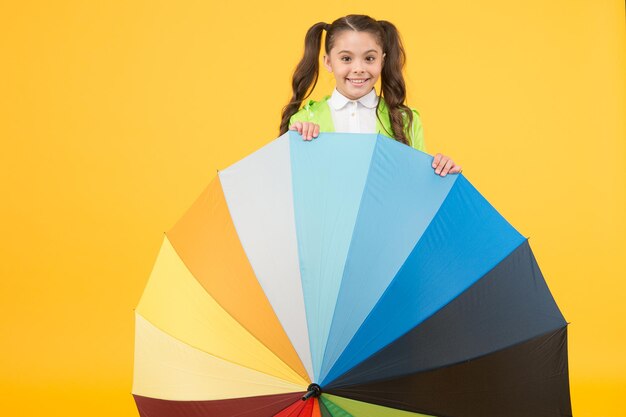 Symbol of protection. Hiding from rain. Smiling schoolgirl peeking out of colorful umbrella. Autumn rain. Going to school rainy days more fun with bright accessories. Water resistant clothes.