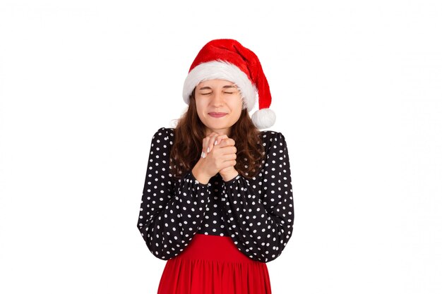 Sympathizing woman in dress holding hands together on chest. emotional girl in santa claus christmas hat isolated 
