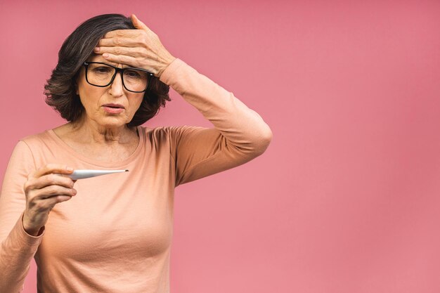 Symptomenconcept van het coronavirus. Rijpe oudere vrouw met griep die elektronische thermometer in de hand houdt, geïsoleerd op een roze achtergrond met kopieerruimte