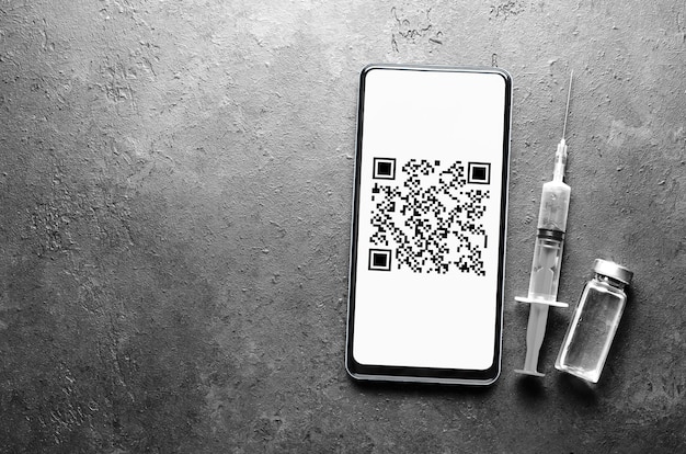 A syringe and a smartphone on a dark background. QR code on the phone screen. The concept of protection and vaccination against viral infection. Flat top view