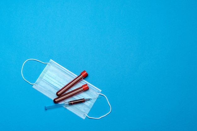 Syringe, test tube with blood sample and disposable face mask to prevent COVID-19 virus on blue