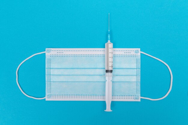 Syringe with medical mask on blue table  flat lay