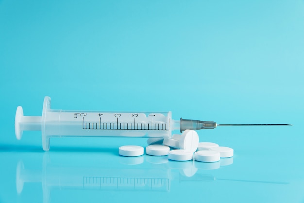 Syringe with needle and white pills scattered over blue colore background. 