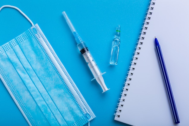 Syringe with vaccine on blue background with a protective face mask and white note for prescription