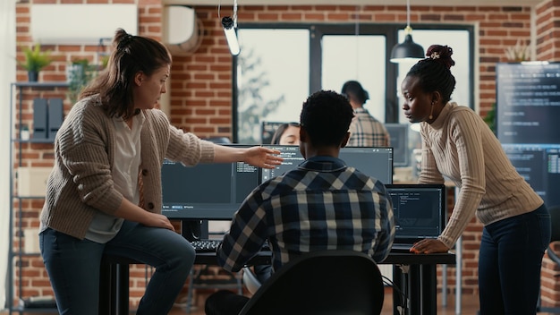 Photo system engineer sitting on desk discussing with mixed team of coders about ai innovation in front of multiple screens compiling code. programmers doing teamwork looking at machine learning algorithms.