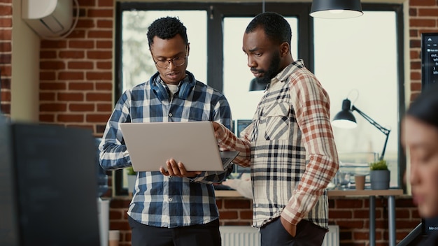 System engineers talking about algorithms holding laptop and standing in the middle of office with colleagues working. Team of software programmers discussing database looking at portable computer.