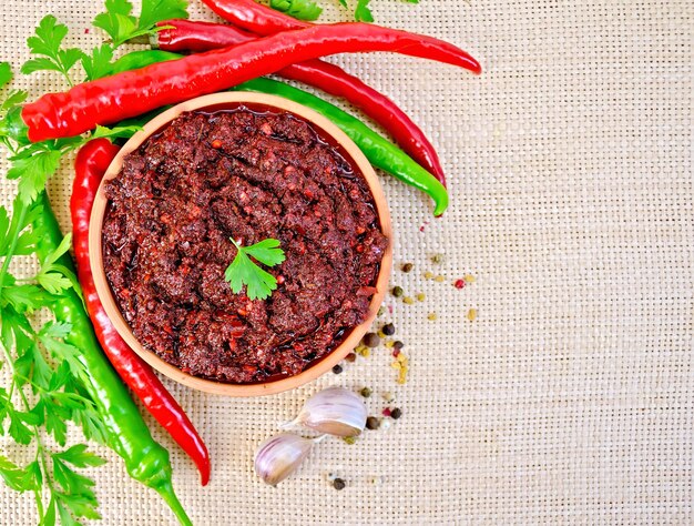 Tabasco adjika in a clay cup, different hot peppers, garlic, parsley on a background of sack cloth