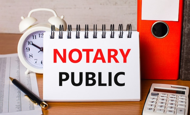 테이블에는 보고서, 흰색 알람 시계, 계산기, 종이 폴더, 펜 및 NOTARY PUBLIC이라는 텍스트가있는 흰색 노트북이 있습니다. 비즈니스 개념