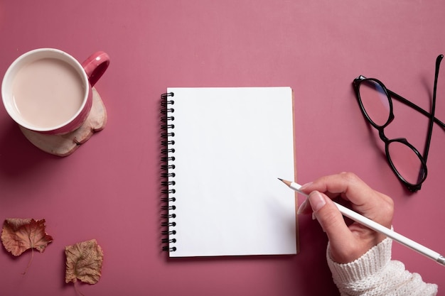 Table desk with notepad and female hand wit pencil and fall leaves on burgundy colored background Autumn flat lay composition with copy space