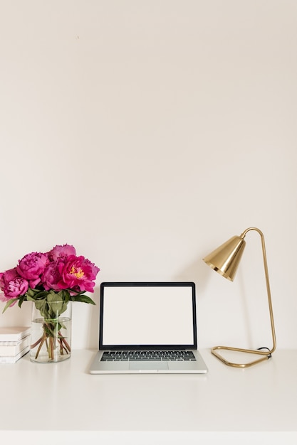 Table desk workspace with blank copy space mock up laptop\
screen. modern home interior design with pink peonies flower\
bouquet, golden lamp against white wall.
