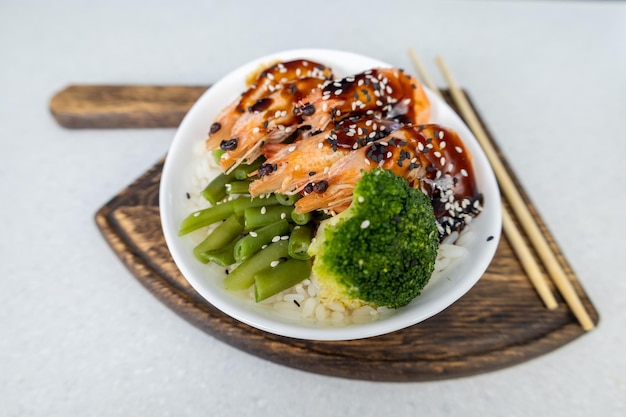 On the table is a plate of rice three king prawns green beans and broccoli on an axeshaped wooden cutting board All topped with unagi sauce Asian food concept