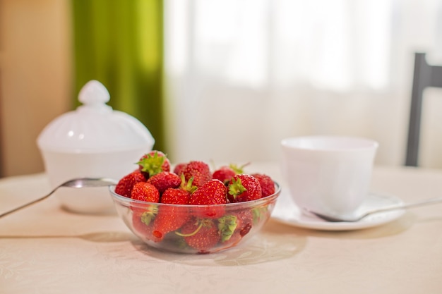 On the table in the kitchen is a hundred of a bowl of strawberries from the home garden. White dishes. Everything is lit by the sun from the window.