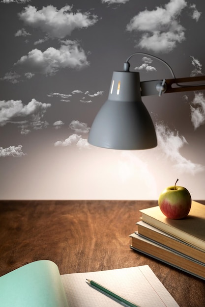 Photo table lamp illuminates the workplace on the office desk a stack of books an open notebook a pencil and an apple on a gray background with clouds