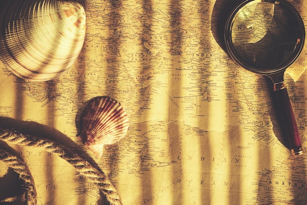 On table, an old map is pinned down with magnifying glass,\
shells, and rope, and sunlight shines through blinds.